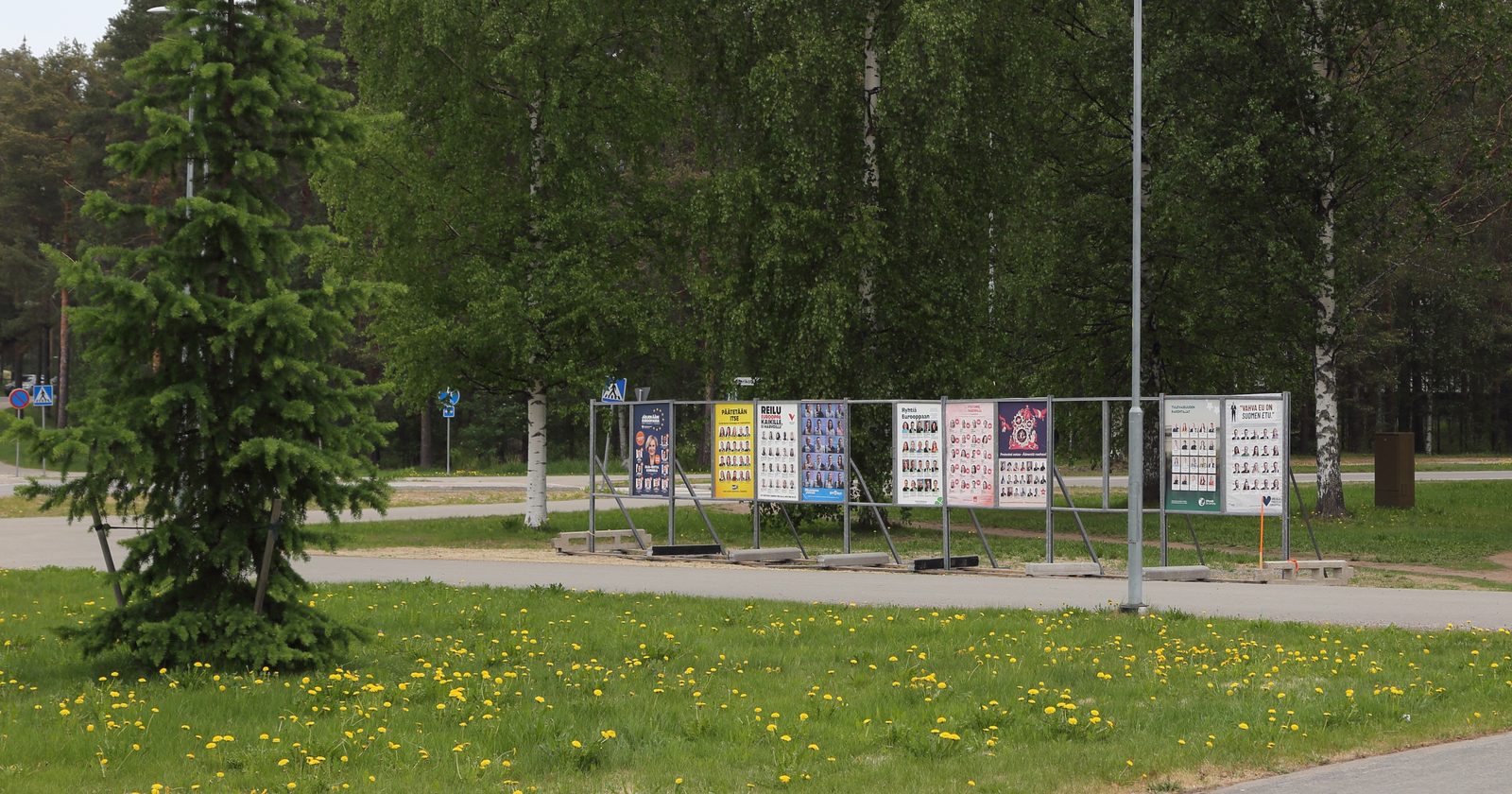 Rząd plakatów wyborczych ustawionych wzdłuż ulicy na osiedlowych tablicach agitacyjnych.