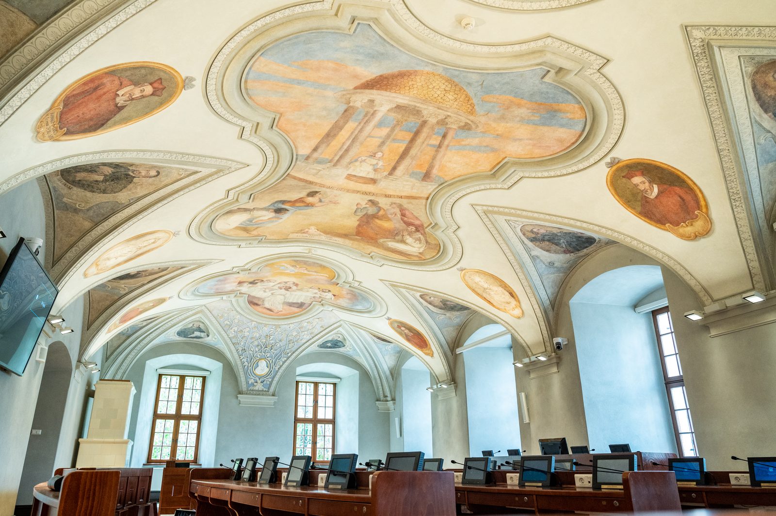 Odrestaurowana sala sesyjna Rady Miasta Poznania z rzędami ław radnych i freskami na ścianach.